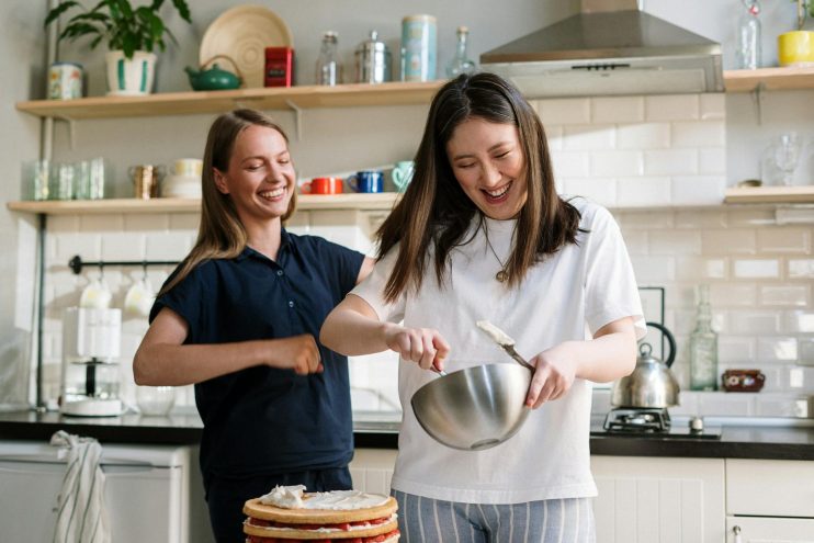 Two women enjoying baking in a modern kitchen, one mixing ingredients in a bowl while the other smiles and encourages her.